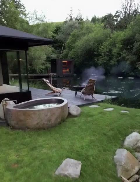 A stone soaking tub and deck chairs beside the pond, woodsmoke drifting